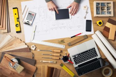 Construction engineer and architect's desk with house projects, laptop, tools and wood swatches top view, male hands using a digital tablet
